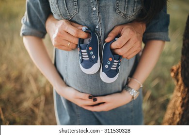 Man And Pregnant Woman Standing On The Road In A Field On A Background Of Rolling Hills And Are Holding Children's Shoes