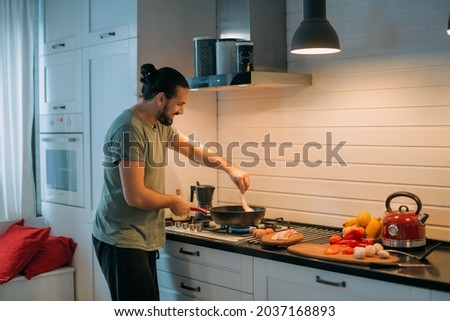 A man prepares breakfast in the kitchen. Young handsome caucasian male preparing food for himself for lunch on a gas stove in a large bright kitchen Foto stock © 