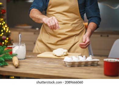 A Man Prepares Food In The Kitchen On Christmas Eve. A Positive Young Adult Man In An Apron Works With Text And Flour. Merry Christmas And Happy Holidays. Decorated With Christmas Lights Interior.