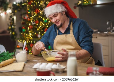 A Man Prepares Food In The Kitchen On Christmas Eve. A Positive Young Adult Man In An Apron And Santa Hat. Merry Christmas And Happy Holidays. Decorated With Christmas Lights Interior.