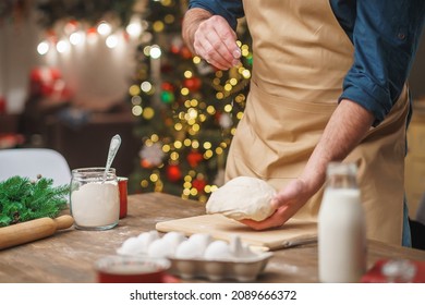 A Man Prepares Food In The Kitchen On Christmas Eve. A Positive Young Adult Man In An Apron Works With Text And Flour. Merry Christmas And Happy Holidays. Decorated With Christmas Lights Interior.