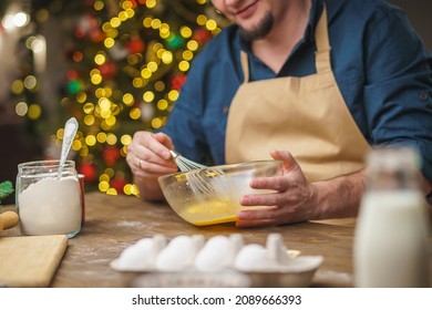 A Man Prepares Food In The Kitchen On Christmas Eve. A Positive Young Adult Man In An Apron And Santa Hat. Merry Christmas And Happy Holidays. Decorated With Christmas Lights Interior.