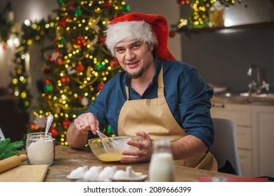 A Man Prepares Food In The Kitchen On Christmas Eve. A Positive Young Adult Man In An Apron And Santa Hat. Merry Christmas And Happy Holidays. Decorated With Christmas Lights Interior.