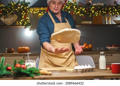 A Man Prepares Food In The Kitchen On Christmas Eve. A Positive Young Adult Man In An Apron And Santa Hat. Merry Christmas And Happy Holidays. Decorated With Christmas Lights Interior.