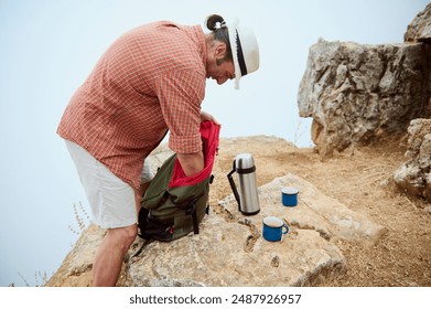 Man preparing coffee while on a camping trip. His backpack and thermos are on a rocky cliff, creating a serene outdoor scene. - Powered by Shutterstock