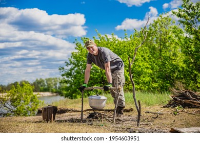 Man preparing fresh fish soup in a pot and dry tree branches for campfire on river bank, resting outdoors