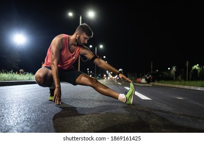 Man Preparing To Run Through The City At Night, He Stretching His Leg Muscles.