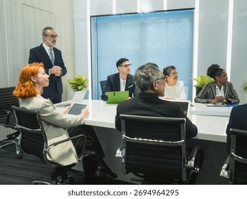 man present plan work to group businessprofessional engaged meeting discuss data display multiple screen show chart graph, use tablet while other seated around conference table with laptop