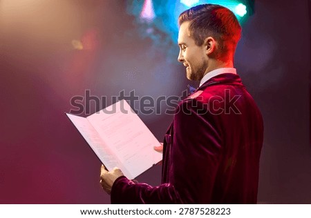 Man presenter host emcee speaker on stage. Showman announcer reads event script under colorful lights, preparing for ceremony, awards and gala. Symbolizes professional hosting and stage performance.