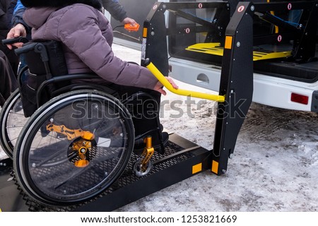 A man presses a button on the control panel to pick up a woman in a wheelchair in a taxi for the disabled. Black lift specialized vehicle for people with disabilities. Yellow handrail. Winter time.