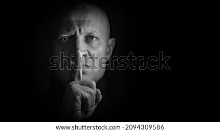 A man presses his finger to his lips and demands to close his mouth, black and white photo. Concept: silence, low key. A mystical phantom in the dark.