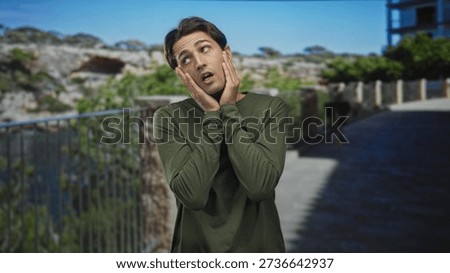 Man pressing hands to cheeks by metal railing on a coastal street walkway, looking upward with worried expression; quiet worry.