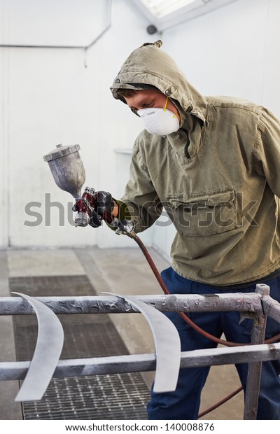 Man in protective clothes and\
respirator paints car details in paint-spraying\
booth