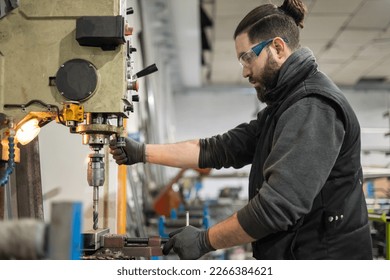 A man in protective clothing who works in a metalworking company is making a hole for a job using a bench drill, or press drill, milling machine. Industrial manufacturing concept. - Shutterstock ID 2266384621