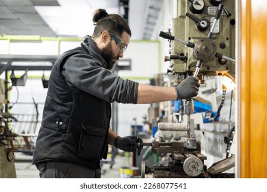 A man in protective clothing who works in a metallurgical company is making a hole for a job using a bench drill, or press drill, milling machine. Concept of small and medium enterprises. - Shutterstock ID 2268077541