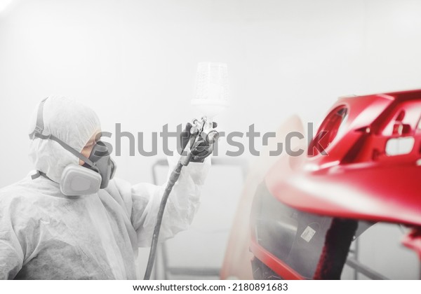 Man in protective mask and\
clothes sprays varnish to car bumper with a spray gun in a paint\
booth.