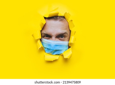 A Man In A Protective Medical Mask Looks In Surprise And Angrily With Curiosity Into A Broken Hole In Yellow Paper. The Concept Of Pandemic, Quarantine, Protection Against Coronavirus And Covid-19.