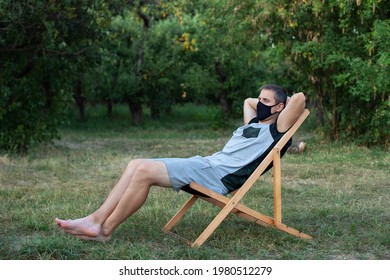 Man In Protective Medical Mask On His Face Sunbathing Outside Green Lawn In Park. Person Outdoors Relaxing On Deck Chair In Garden. Guy On Lounge Sunbed On Summer Weekend. Person Enjoy Rest Outdoors. 