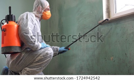 A man in a protective suit, glasses and a respirator sprays a disinfectant. Mold remediation specialist in uniform inspects walls and spraying pesticide on damaged wall with sprayer