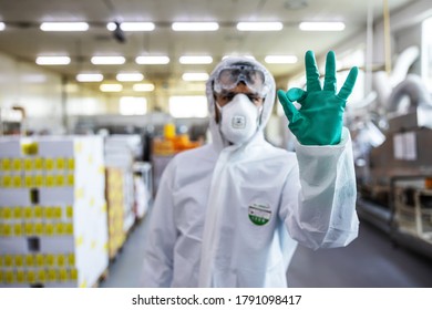 Man In Protective Suit, Mask And Gloves Standing In Food Production Factory And Showing Okay Sign. He Just Disinfected Whole Facility From Covid-19 / Corona Virus.