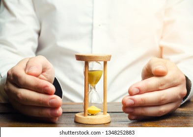 Man Protects The Hourglass. Concept Of Saving Time And Money. Time Management. Planning Work. Reduced Cost And Bureaucratic Burden. Saving Productivity. Extension Of Life. Debt / Tax Holidays