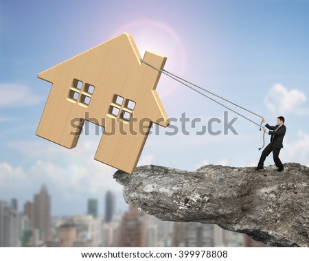 Man pulling rope to move wooden house on cliff edge, with sun sky cityscape background.