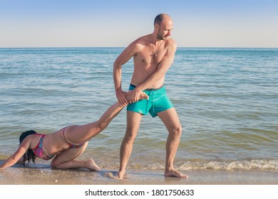 A man pulls a woman by the leg along the sea coast. The vacation is over, it's time to go home. The husband picks up his wife from the beach.