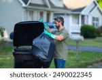 A man pushes a large plastic trash bin for weekly waste disposal schedule. Middle aged man putting out rubbish in garbage bin at the street. Man throwing trash bag into bin outdoors.