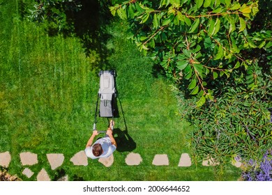 Man Pushing Lawn Mower For Cutting Green Grass In Garden With Sunlight At Summer Season. Aerial View. Housework And Lifestyle Concept. 