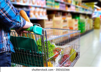 Man Pushing Shopping Cart Full Of Food In The Supermarket Aisle. Elevated Rear View. Horizontal Composition