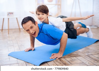 The Man Pushup From The Floor With The Boy On His Back. This Is Father And Son. They Are At Home.