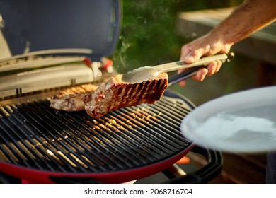 Man Put Down On A Wooden Board Ready To Eat Grilled Steak Meat, Male Roasted Steak Meat On The Gas Grill On Barbecue Grill Outdoor In The Backyard, Summer Family Picnic, Food On The Nature.