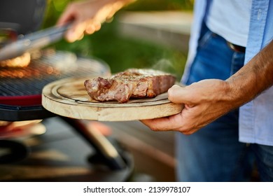 Man Put Down On A Wooden Board Ready To Eat Grilled Steak Meat, Male Roasted Steak Meat On The Gas Grill On Barbecue Grill Outdoor In The Backyard, Summer Family Picnic, Food On The Nature.