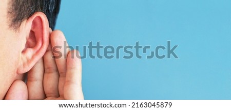 Man put his hand to his ear and listens carefully on blue background copy space.