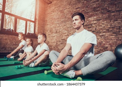 Man Put His Legs Together And Holds Them With His Hands. Young Family Sitting On A Mat In The Gym Perform Muscle Building Exercise. 