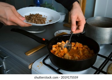 Man Puts Buckwheat And Chicken From A Pot And Frying Pan On A Plate, Hands Closeup. Healthy Food Of Poor Person In Third World Country. Old Time Kitchen Utensils Pot And Frying Pan On The Gas Stove.