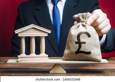 A Man Puts A Money Bag On The Scales Opposite To The Building Of The Government, Bank, University. Budget And Funding For Normal Functioning. Payment Of Taxes. Deposits And Loans. Lobbying Interests.