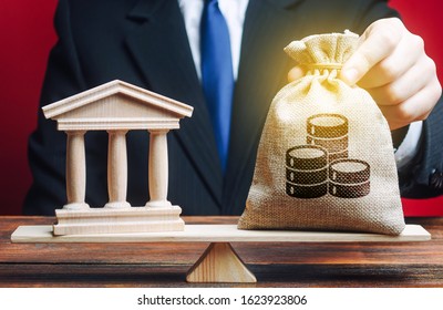 A Man Puts A Money Bag On Scales Opposite To Building Of Government, Bank, University. Payment Of Taxes. Deposits And Loans. Budget And Funding For Normal Functioning. Lobbying Interests.