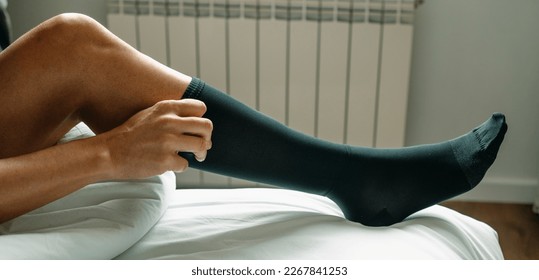 a man puts on a black compression sock sitting on his bed at home, in a panoramic format to use as web banner or header