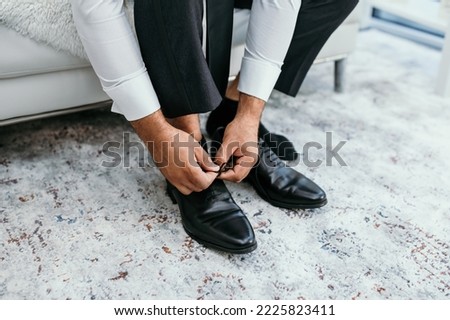 A man puts on black shoes in a hotel room. A man in a white shirt is tying shoelaces on leather shoes close-up. Businessman about to get dressed and going to a business meeting