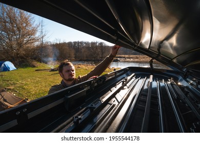 A Man Puts Things In The Roof Rack Of A Car Or In A Cargo Box, On A Blurred Background Of A River And Trees, On A Summer Day. Family Trip. Close-up Inside View.