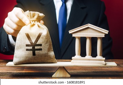 A Man Puts A Yen Yuan Money Bag On Scales Opposite To Building Of Government, Bank, University. Budget And Funding For Normal Functioning. Deposits And Loans. Lobbying Interests. Payment Of Taxes.