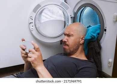 A man putting dirty laundry into washing machine and lie playing on the phone.