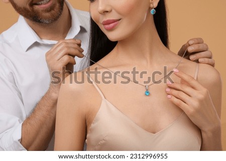 Man putting elegant necklace on beautiful woman against beige background, closeup