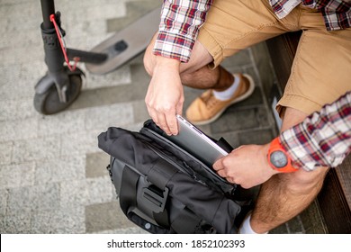 Man Putting Laptop In Backpack Outside Sitting Near Electric Transport Scooter. Caucasian Man Packing Laptop In Backpack. Male Pulling Laptop Out Backpack. Person With Computer Sitting On Wooden Bench