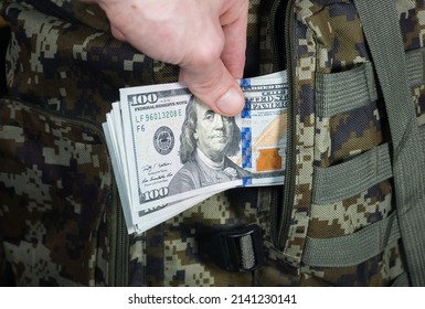 Man Is Putting In A Military Backpack Dollars. Dark Green Camouflage Khaki Bag With Money. Migrants And Refugees Concept.