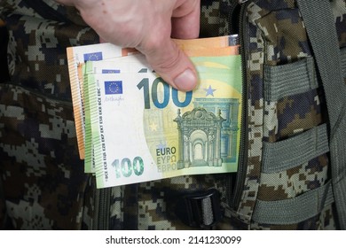 Man Is Putting In A Military Backpack Euros. Dark Green Camouflage Khaki Bag With Money. Migrants And Refugees Concept.