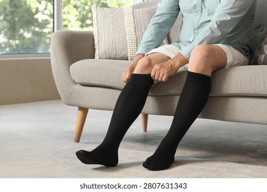 Man putting on black compression socks while sitting on sofa