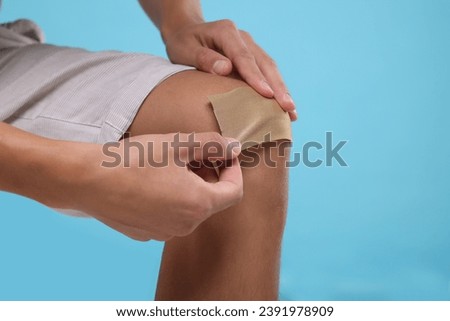 Man putting sticking plaster onto knee on light blue background, closeup. Space for text
