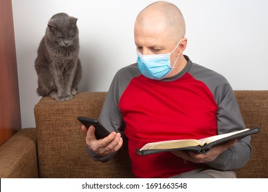 Man In A Quarantine Period In A Protective Mask Studies A Bible Using A Smartphone Next To A Gray Cat. The Study Of Religion And Christianity